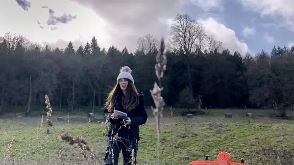 A female researcher stands in a field beside an orange drone, preparing to start a flight. She is wearing a bobble hat.