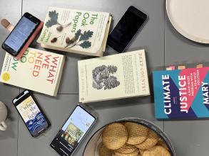 Books and phones are scattered across a grey kitchen table, next to a biscuit tin and a cup of tea